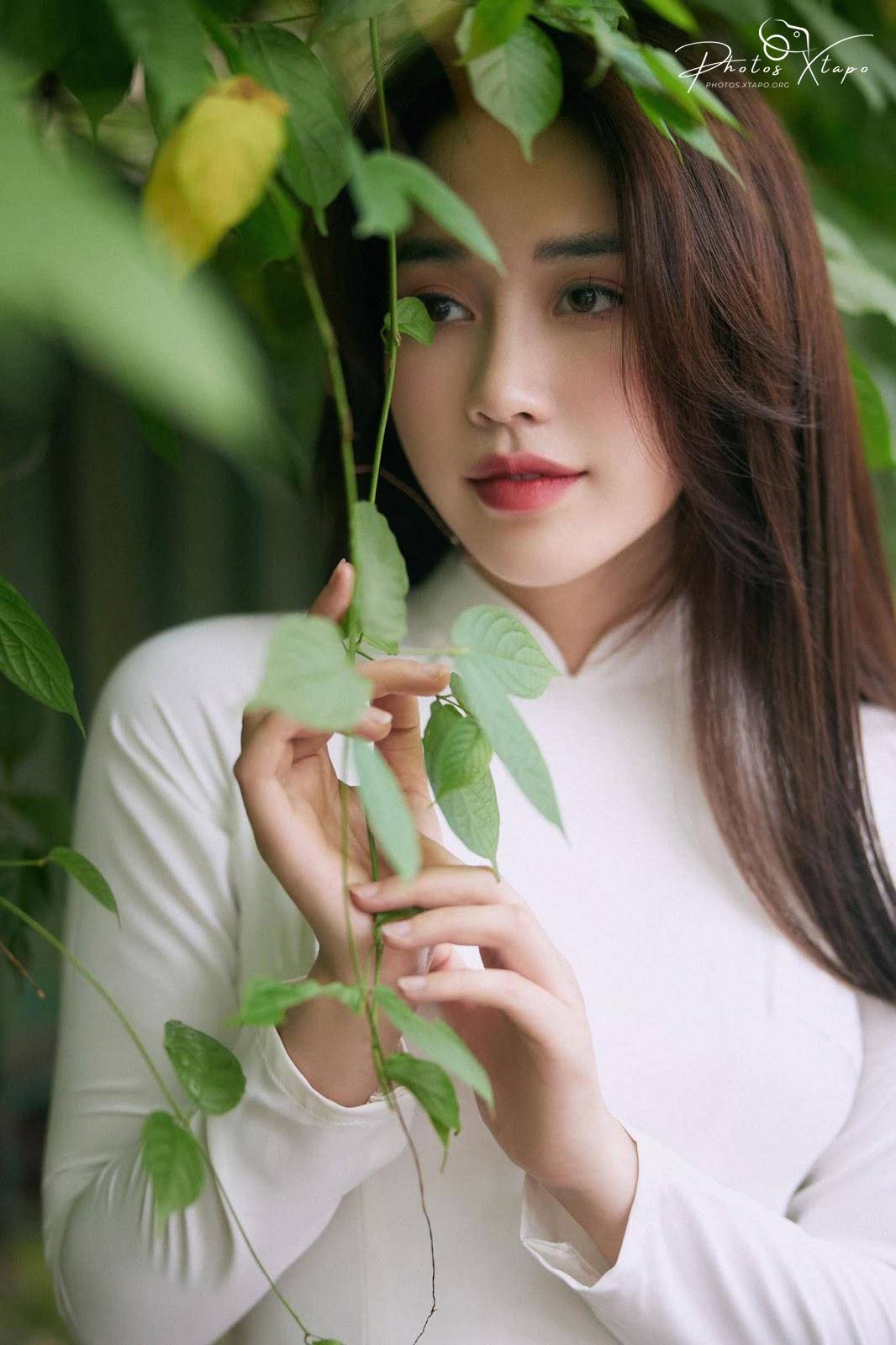 A young Vietnamese woman in a traditional white ao dai poses amidst lush green foliage.