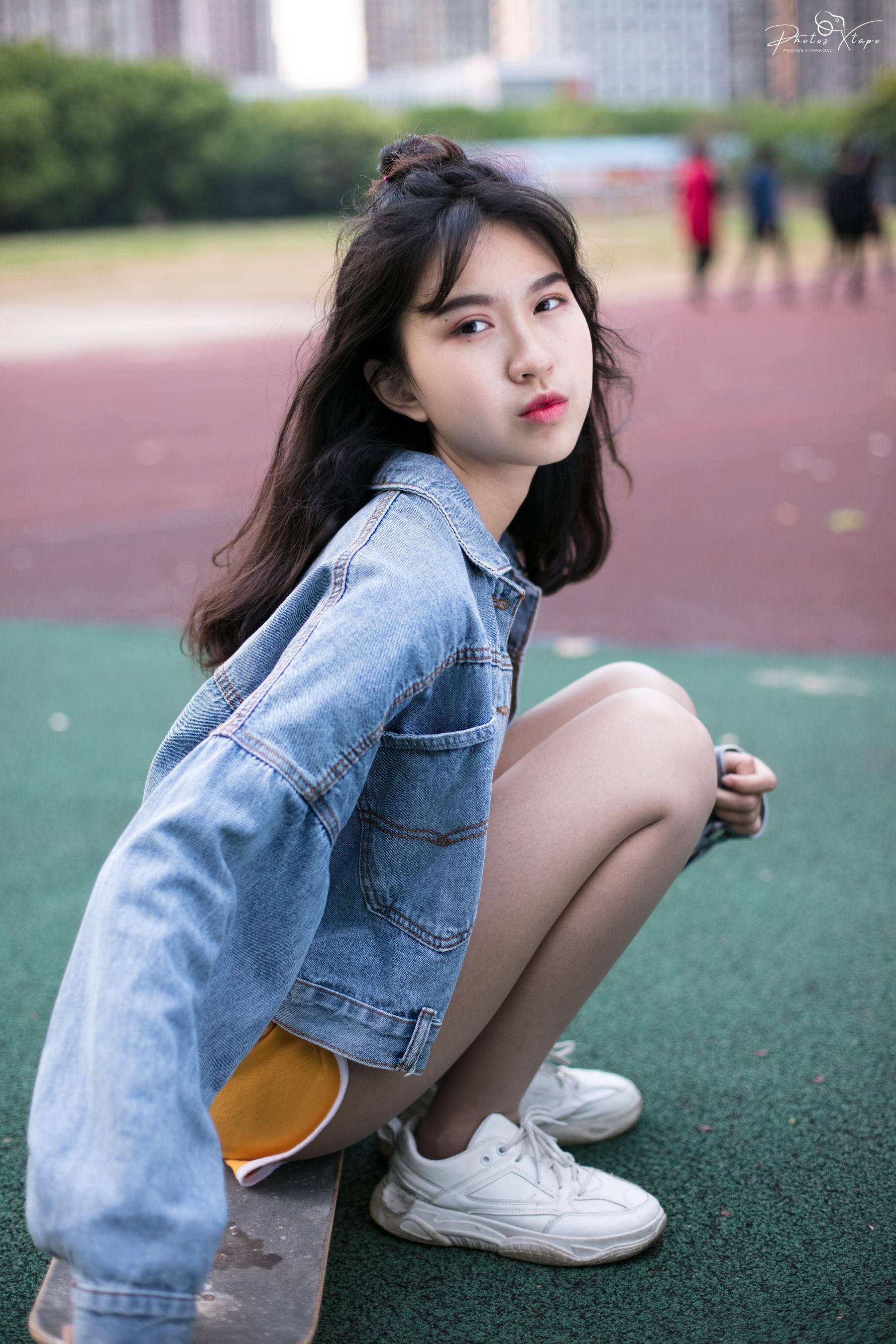 A young woman sits on a skateboard, looking over her shoulder. She's wearing a denim jacket and white sneakers.