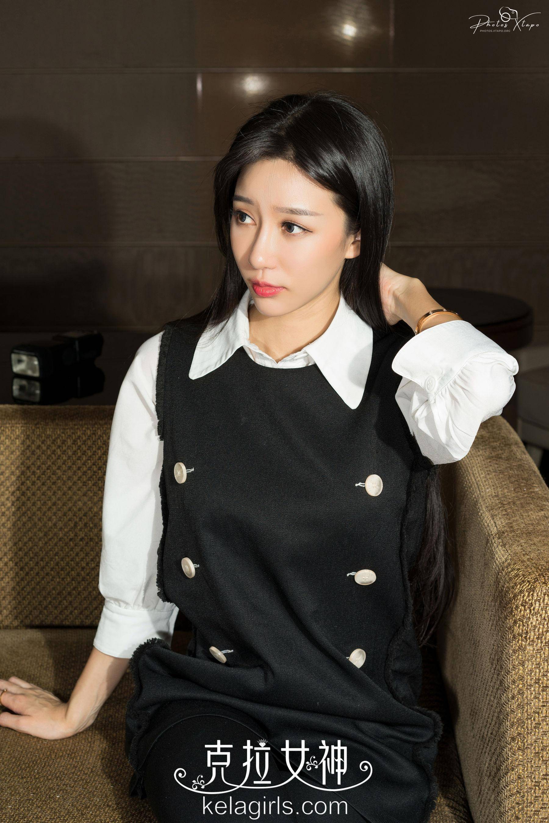 A beautiful young woman sits elegantly on a couch, wearing a stylish black dress and a crisp white shirt. Her long, dark hair cascades down her shoulders. The image exudes sophistication and grace.