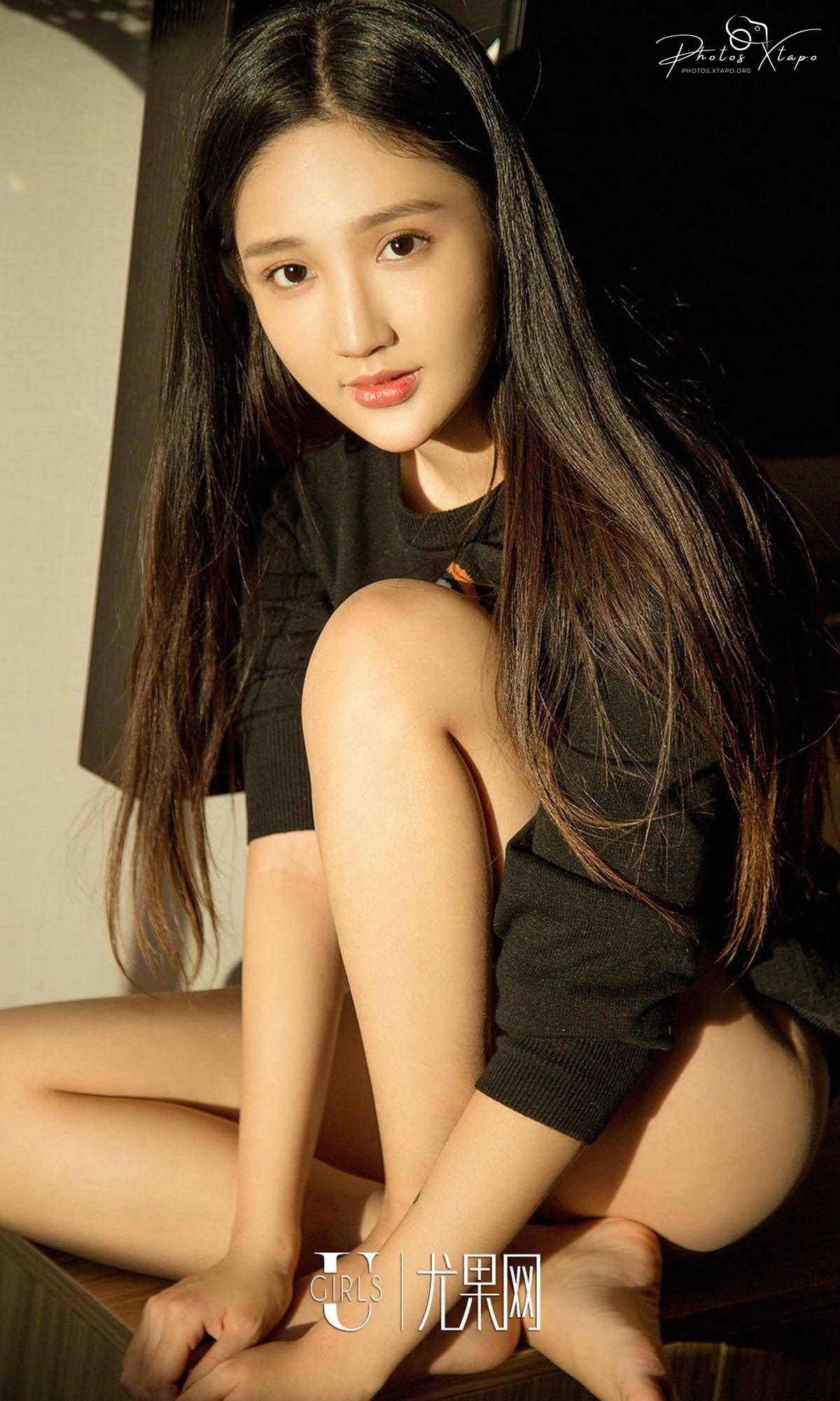 A young Asian woman with long dark hair sits on a surface, her legs crossed, wearing a black sweater. Soft, warm lighting enhances her features.