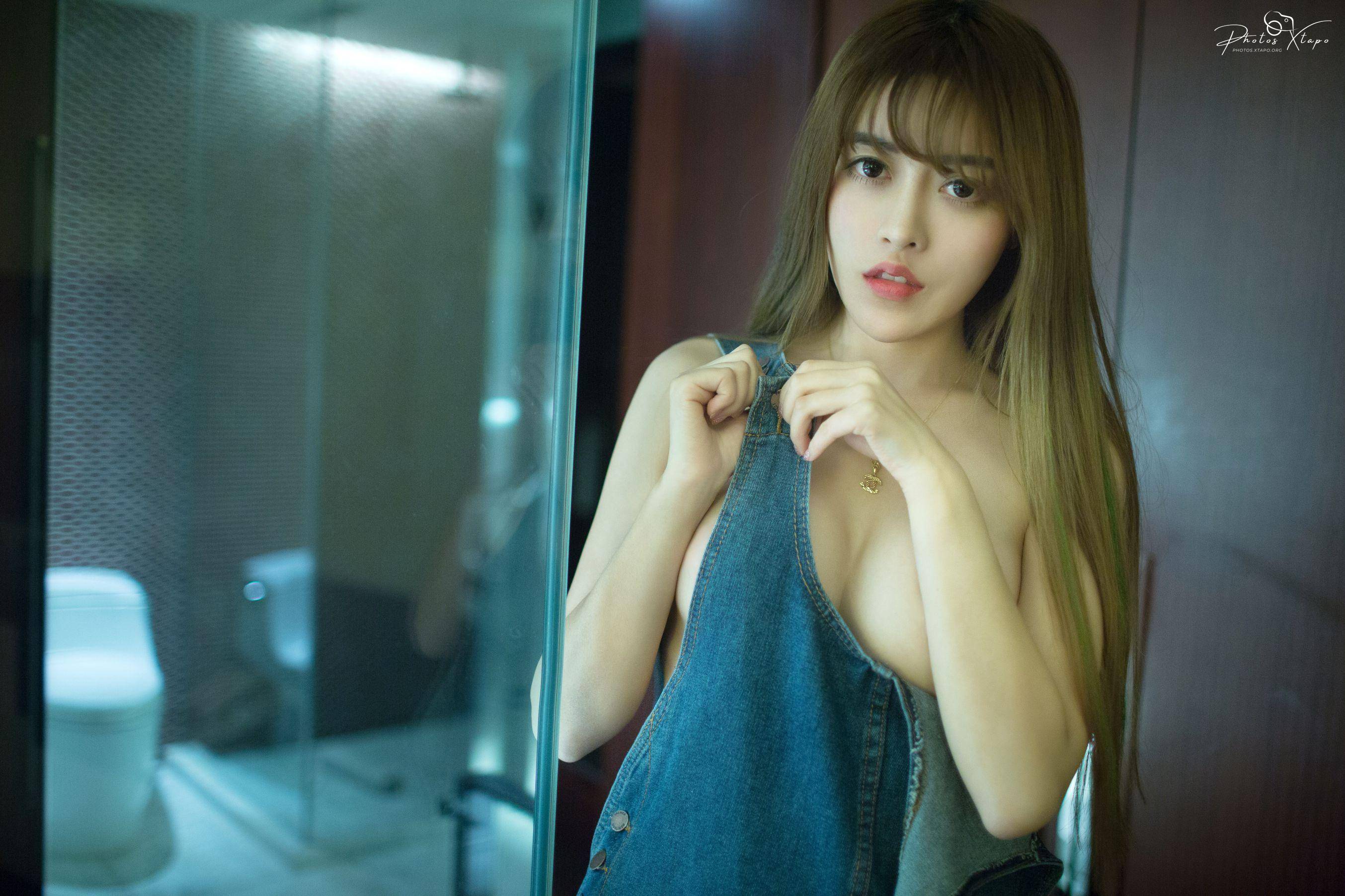 A captivating portrait of a young Asian woman, partially revealing her upper body, while she playfully adjusts her denim overalls. The image exudes allure and confidence.