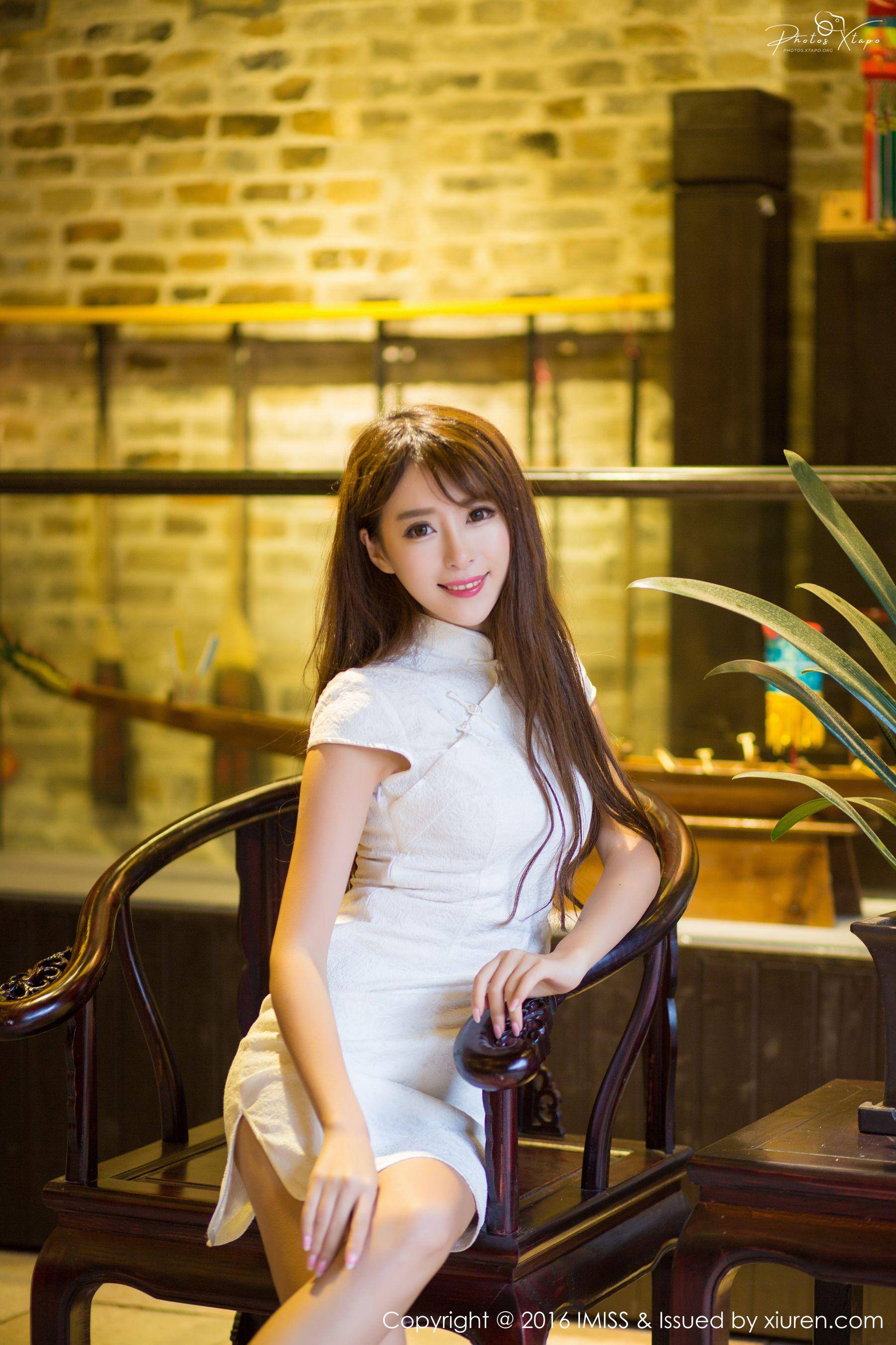 A young woman in a white cheongsam sits gracefully on an antique chair, posing against a backdrop of a rustic brick wall.
