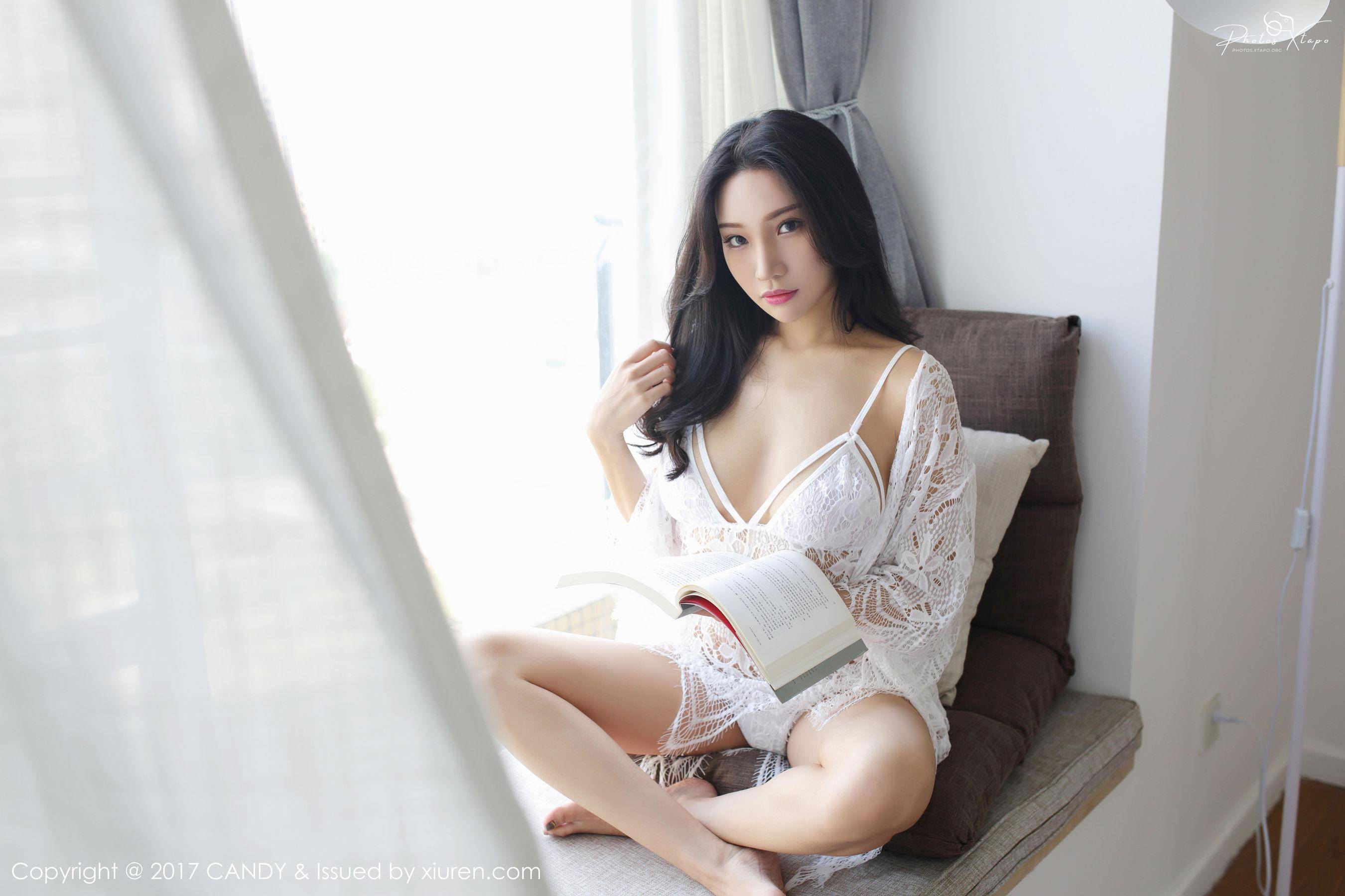 A young woman sits by a window in a white lace robe and holds a book.