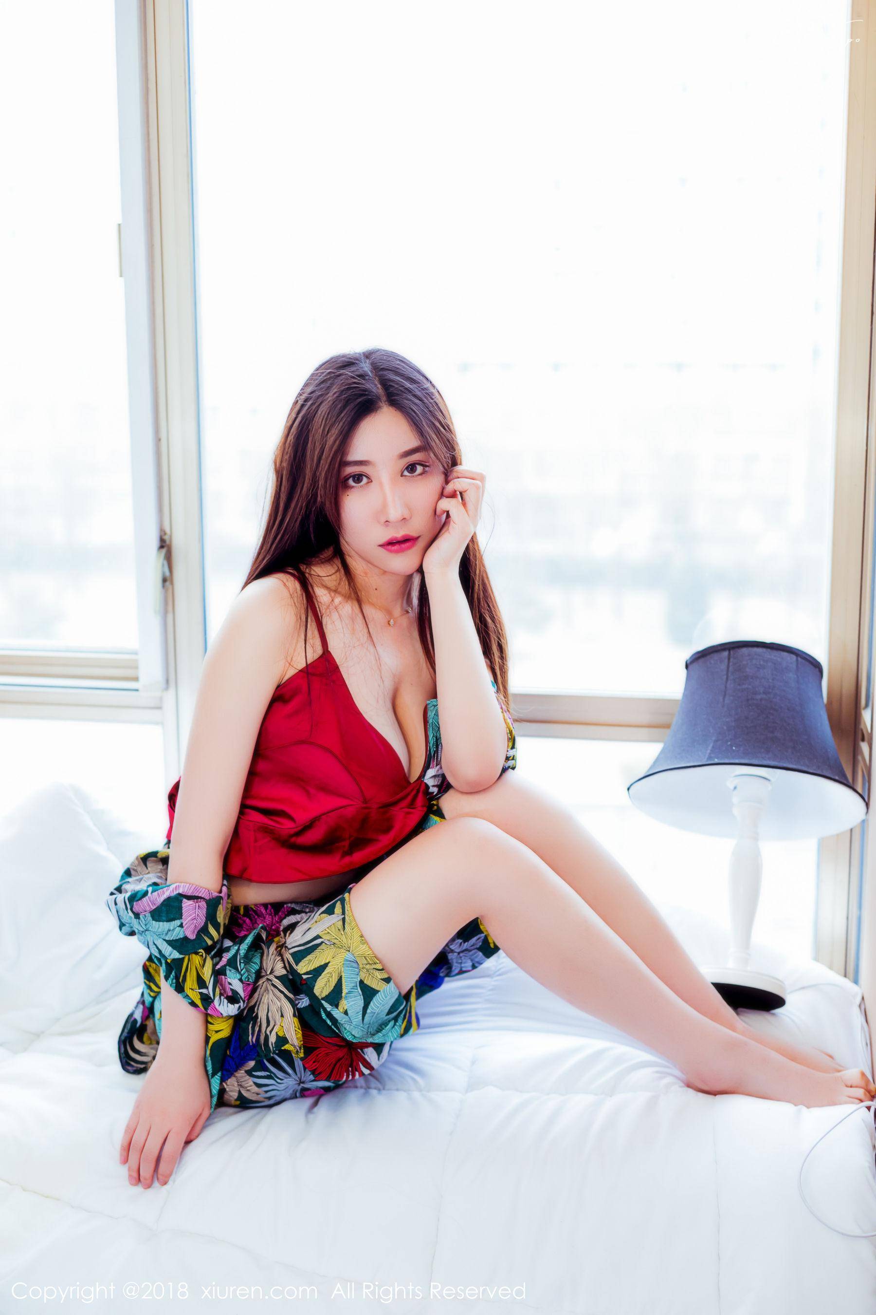 A young woman with long brown hair sits on a bed, dressed in a red satin top and floral shorts. She is looking at the camera and has one hand on her face.