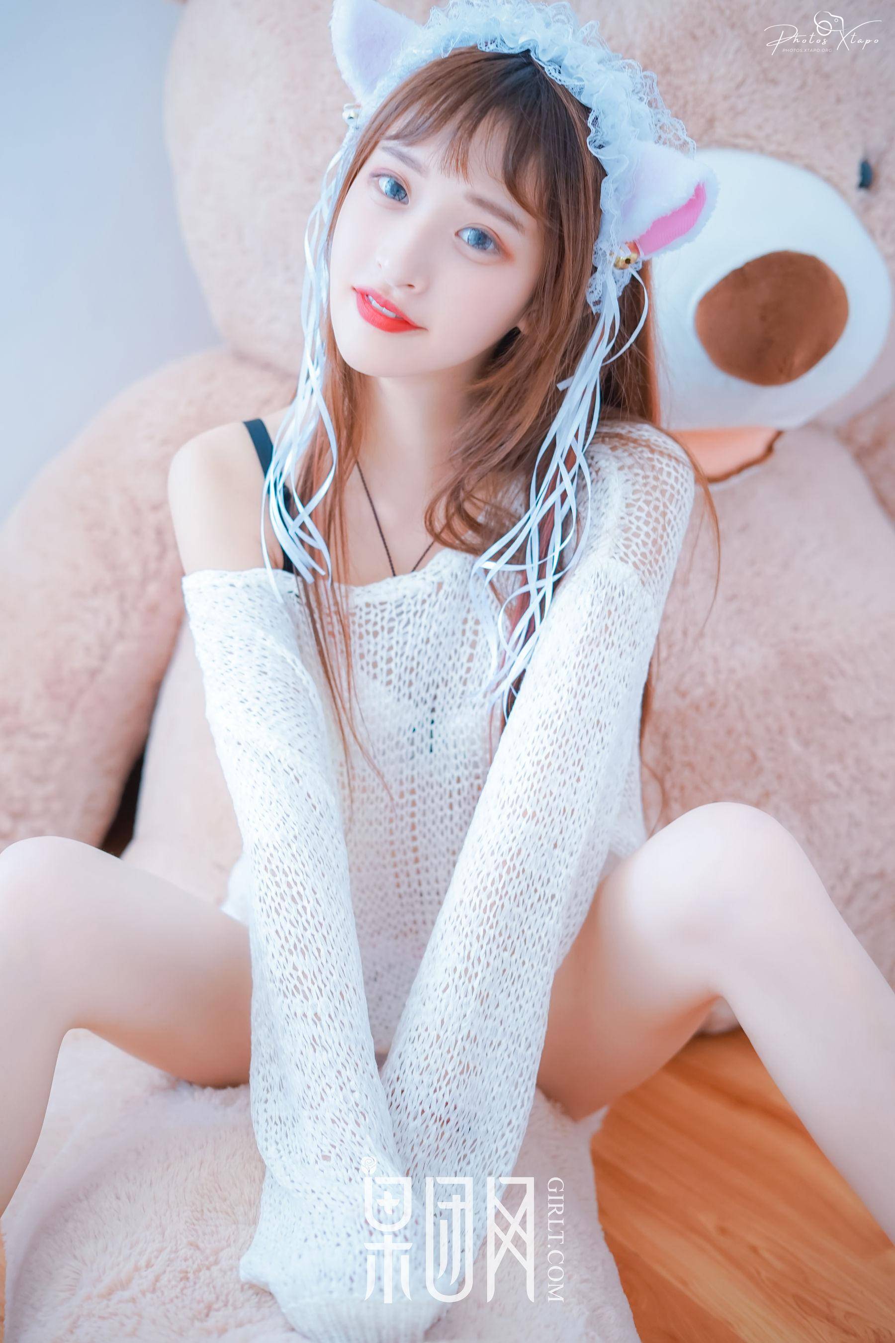 A young woman wearing a white knit sweater and a cute cat-ear headband sits on a plush teddy bear.  She has long brown hair and is looking directly at the camera.