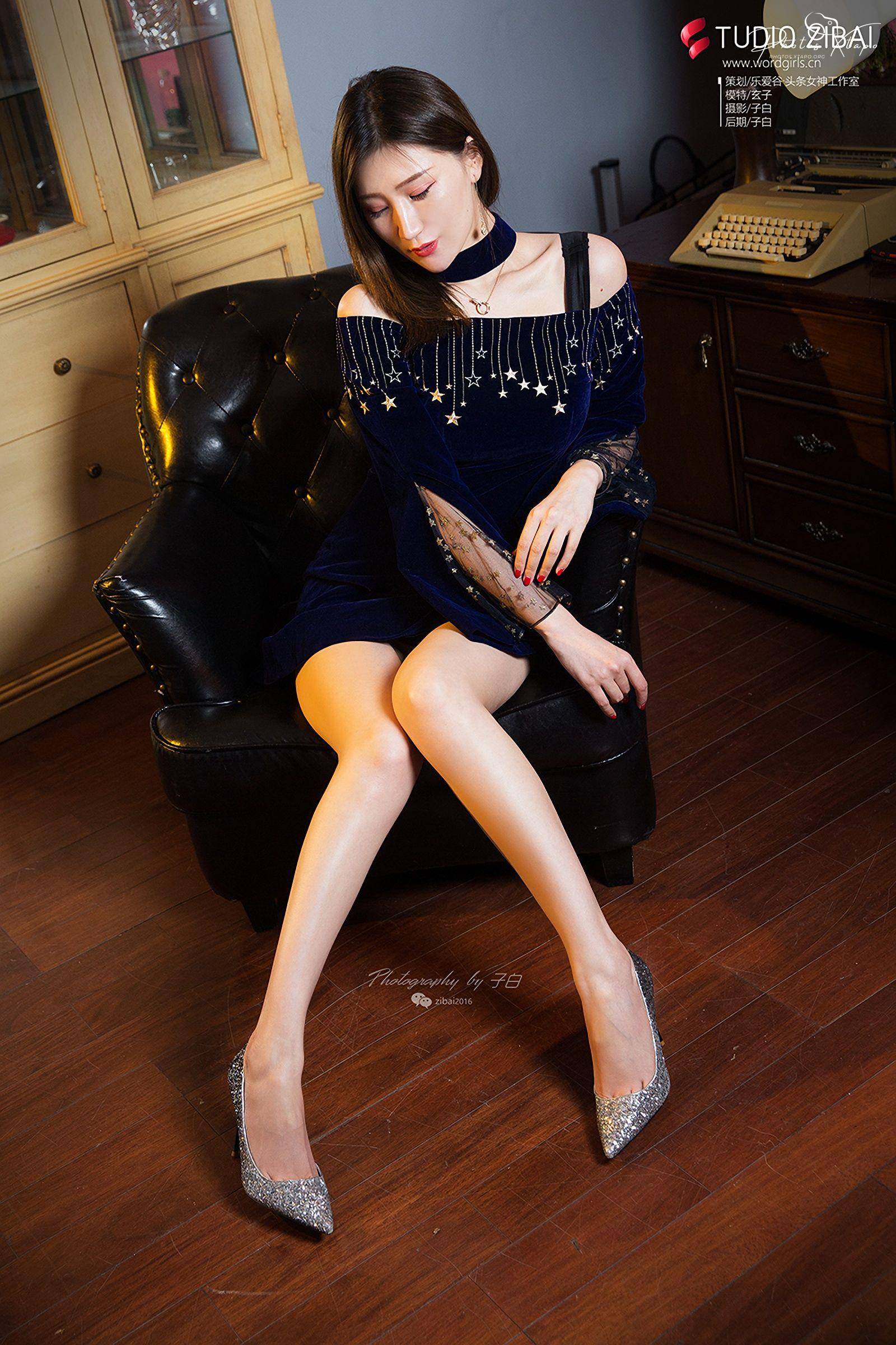 A woman in a navy blue velvet dress adorned with stars sits in a plush armchair. She wears silver glitter heels and a delicate necklace.