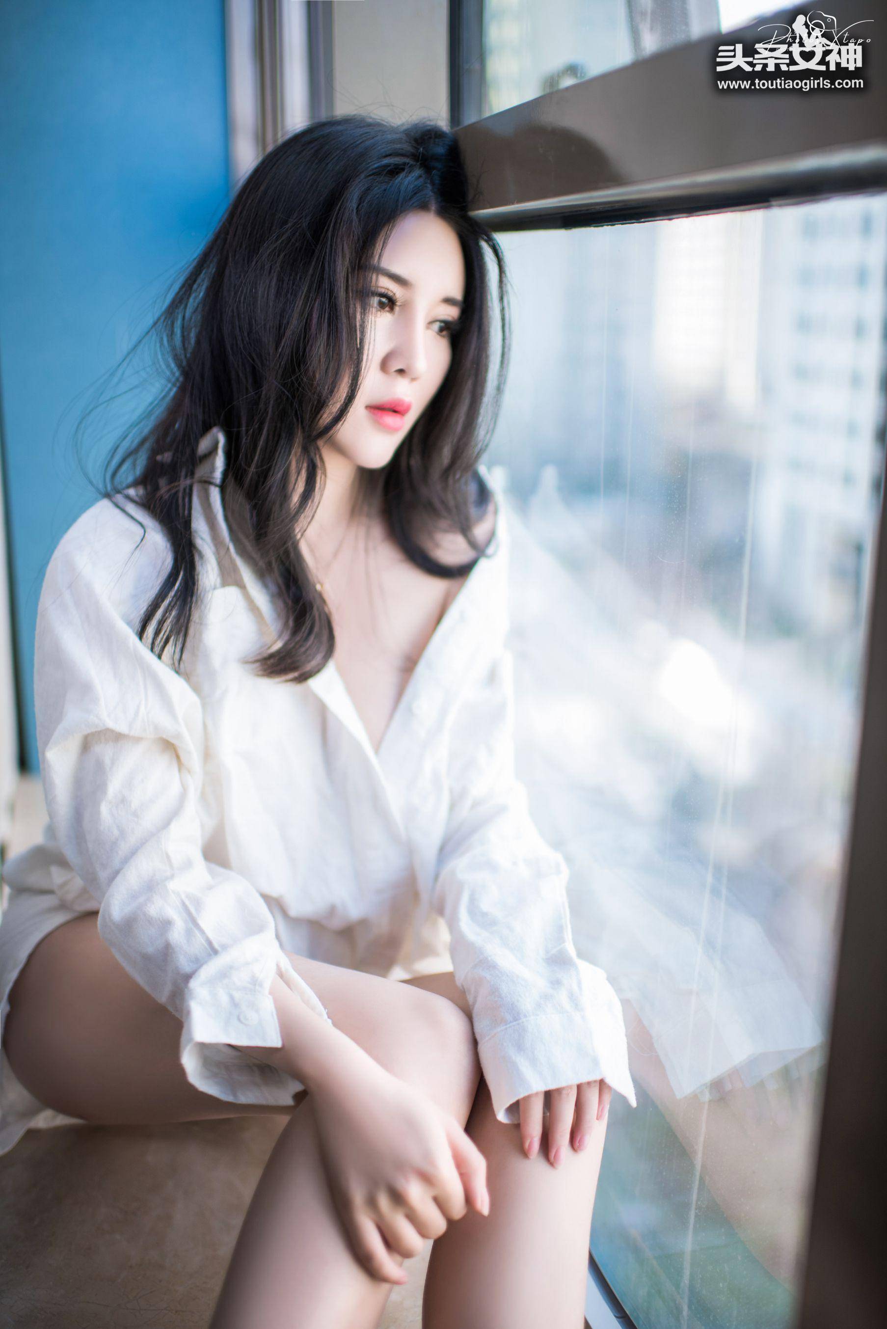 A young woman with long black hair sits by a window wearing a white shirt.