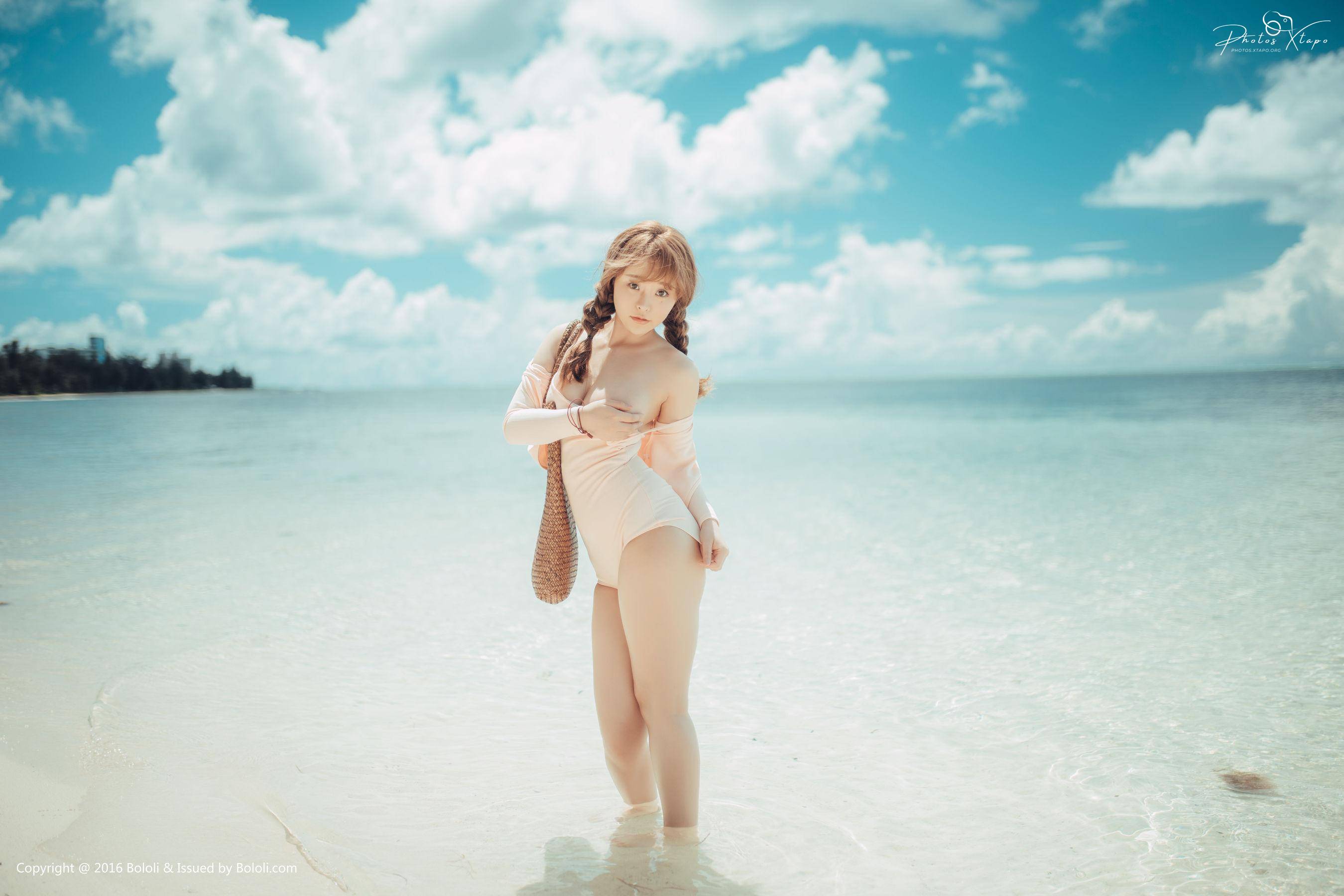A young woman in a light pink swimsuit poses on a beautiful beach with crystal-clear water and a bright sunny sky.