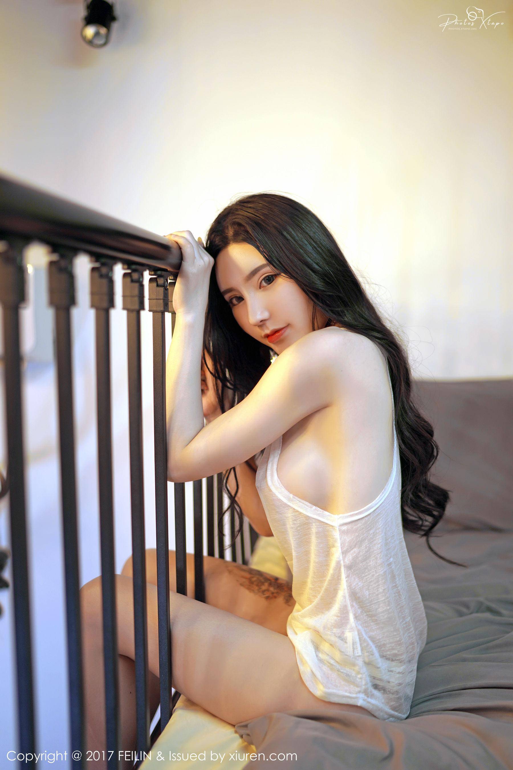 A young woman with long black hair poses in a white tank top, leaning against a railing. Her skin is fair, and she looks directly at the camera.